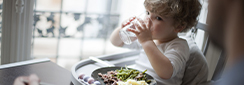 Un jeune enfant boit son verre d'eau devant son assiette, assis dans sa chaise bébé